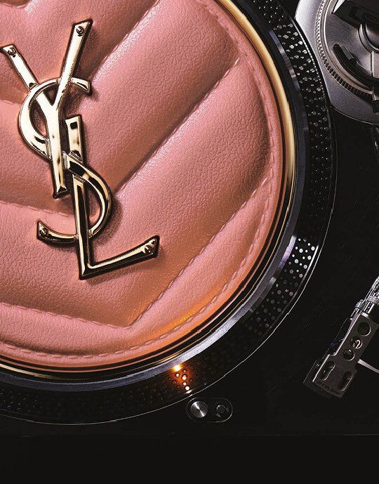 Gros plan d'un compact ou accessoire Yves Saint Laurent rose matelasse orne du logo YSL dore en son centre. L'objet est pose sur le bord texture metallique d'une platine vinyle, fusionnant le luxe de la beaute avec une esthetique inspiree de la musique.