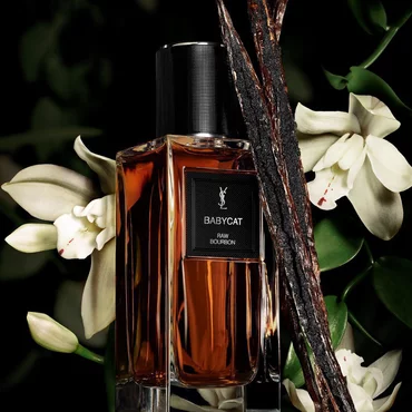 Une bouteille de parfum couleur ambre etiquetee "BABYCAT RAW BOURBON" de YSL, ornee de gousses de vanille et de fleurs d'orchidees blanches.