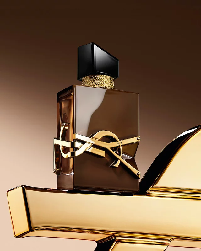 Flacon Libre Santal Couture brun glace refletant la lumiere, pose sur une grande Cassandre YSL doree.