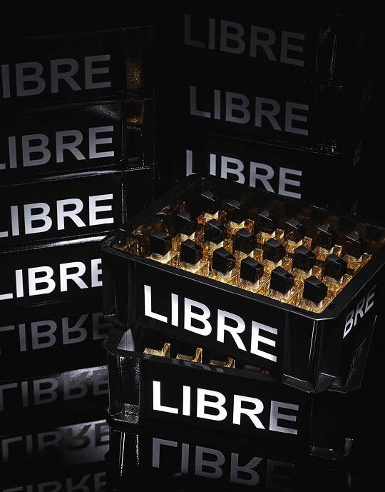 Une composition de caisses noires, semblables a celles utilisees pour le transport de bouteilles, empilees et remplies de flacons du parfum YSL Libre. Les caisses portent l'inscription "LIBRE" en grandes lettres blanches lumineuses. Au premier plan, une caisse est remplie de multiples flacons du parfum "Libre" qui refletent la lumiere, creant des eclats dores. L'ensemble est baigne dans une lumiere sombre, accentuant le cote chic et la mise en scene du produit.