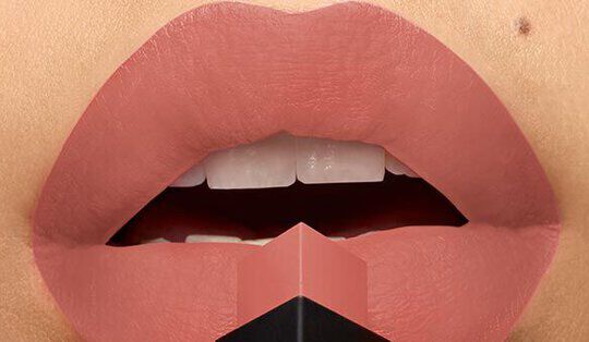 Close-up of a pair of lips with a nude matte lipstick shade, partially open and positioned above a small lipstick bullet.