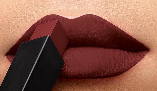 Close-up of a pair of lips with a deep matte lipstick being applied from a black and red lipstick tube.