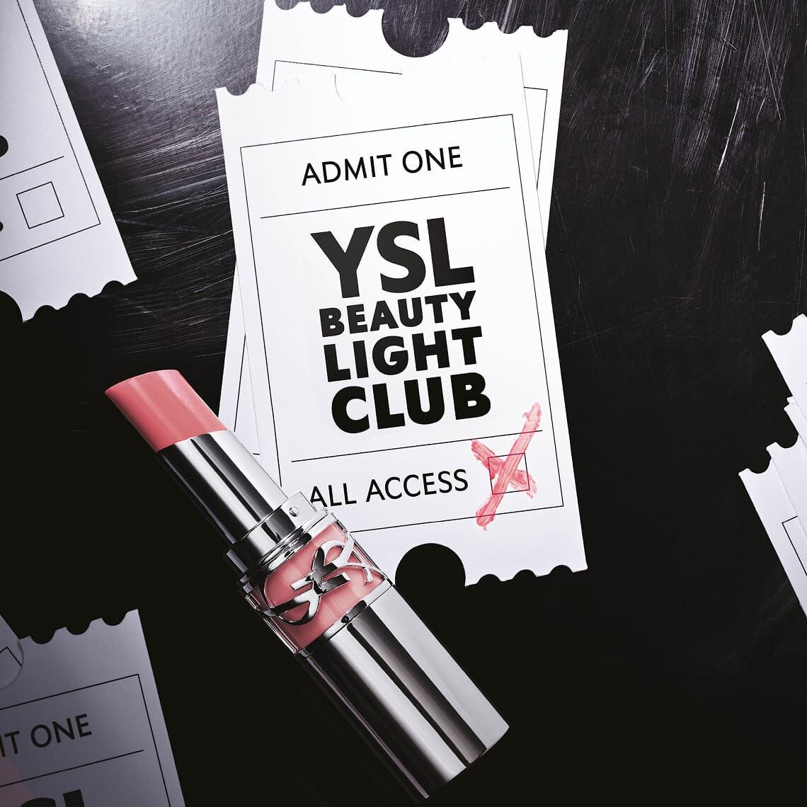 Un rouge a levres rose pale a la texture brillante est pose sur deux tickets blancs de type evenementiel, legerement superposes. Le ticket superieur porte le texte "ADMIT ONE" en haut, et au centre, "YSL BEAUTY LIGHT CLUB" en grandes lettres noires, avec "ALL ACCESS" en dessous. Une croix rouge est griffonnee sur le coin inferieur droit du ticket, comme pour le valider. L'arriere-plan est sombre, evoquant une surface texturee, et on apercoit des bords de tickets dechiquetes sur les cotes, suggerant plusieurs autres tickets disperses. L'ensemble donne une impression d'invitation exclusive a un evenement glamour.