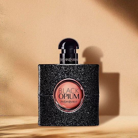 Un flacon de parfum Black Opium avec une finition noire pailletee et une etiquette ronde rose affichant "BLACK OPIUM YVES SAINT LAURENT" au centre. Le flacon est muni d'un bouchon sombre et repose sur une surface en bois sur un fond beige clair avec des ombres.