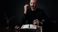 Un parfumeur en costume sombre sent attentivement une touche a parfum dans un cadre faiblement eclaire.