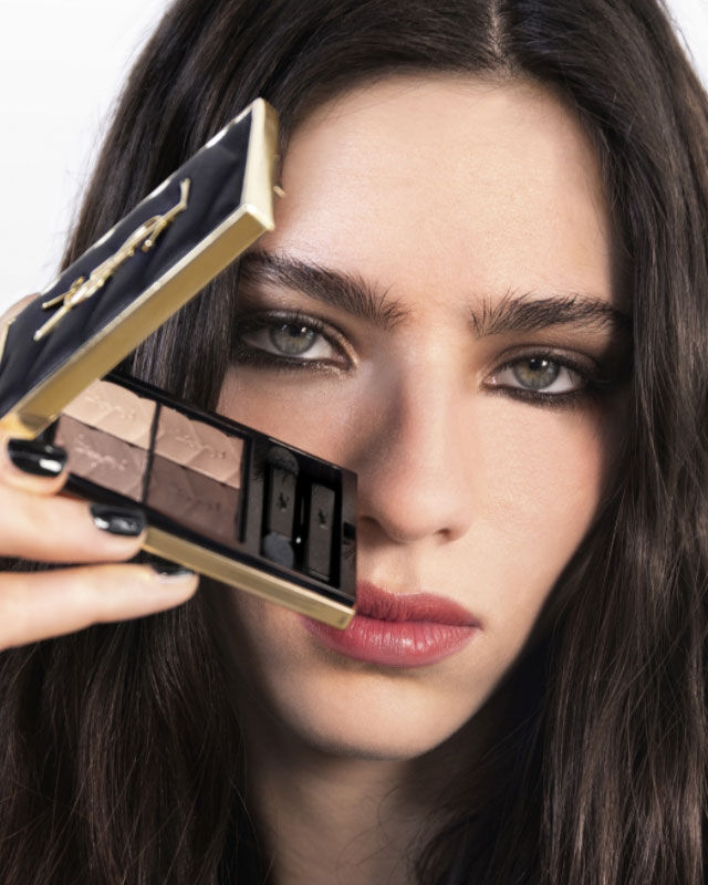 Femme avec de longs cheveux fonces tenant une palette de maquillage pres de son visage, affichant un look sophistique avec des yeux charbonneux.