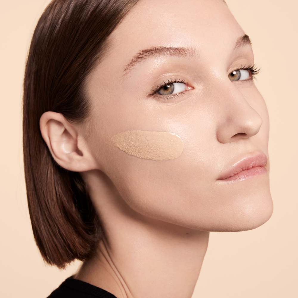 A model with a neutral expression is shown with a swipe of foundation on her cheek, showcasing a natural makeup look against a soft beige background.
