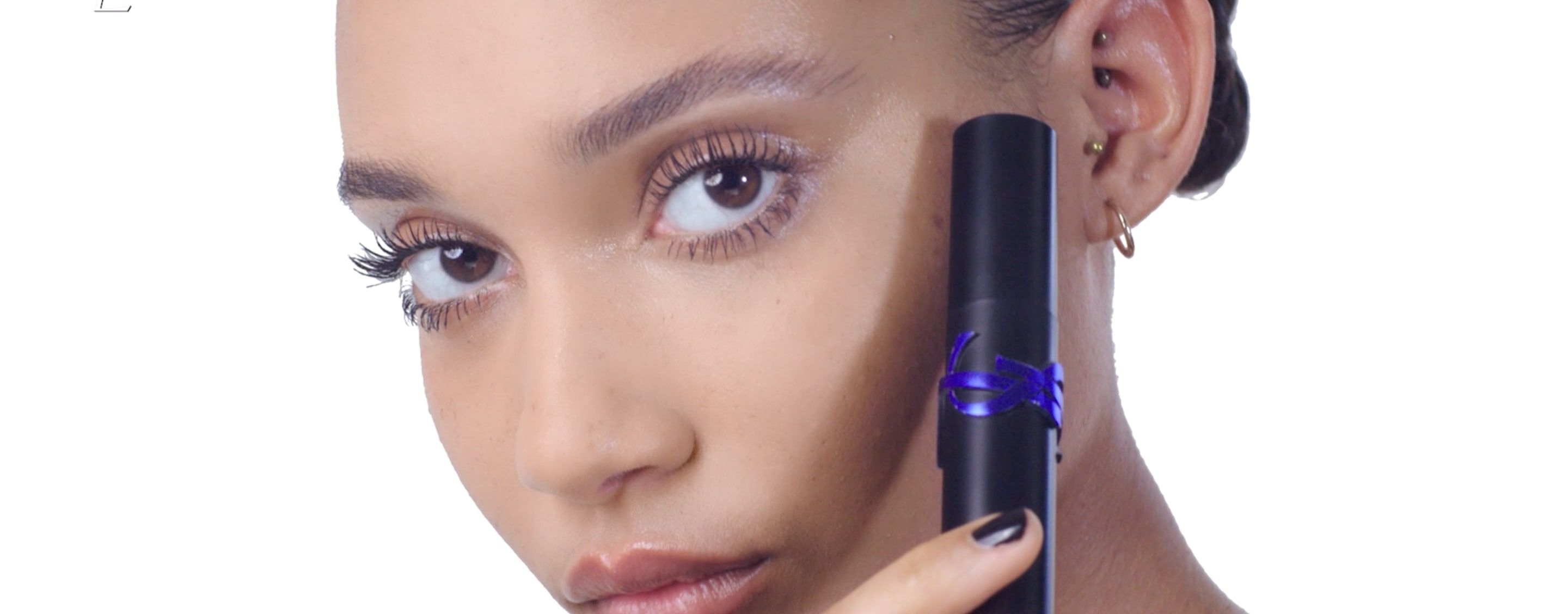 A close-up of a person with long eyelashes holding a black cosmetic product close to their face, with a neutral background.