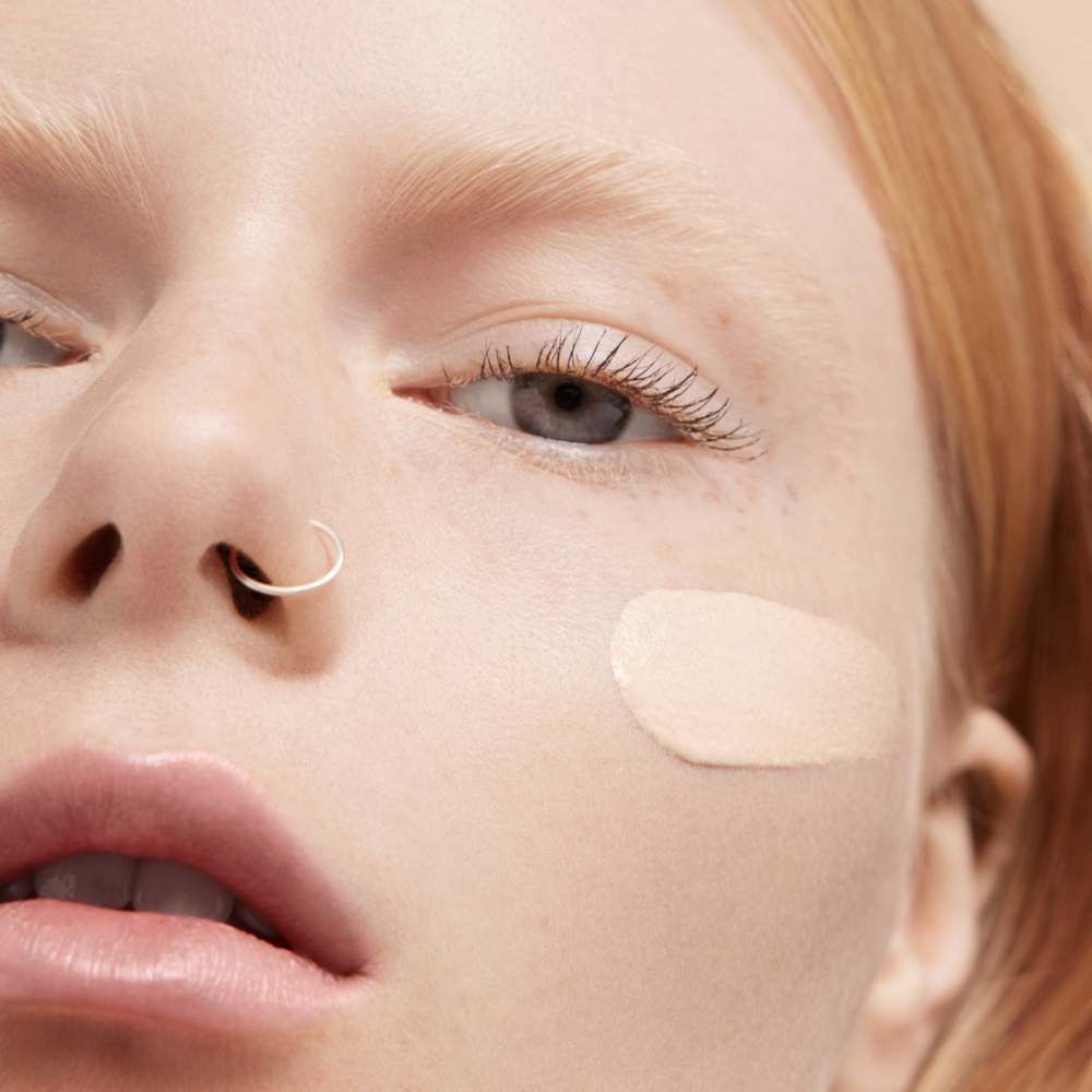 A close-up of a person's face with light skin and red hair, featuring a band of foundation applied to one cheek. The person's expression is neutral, and they have a nose ring.