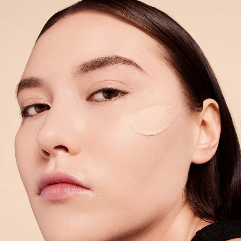 A close-up of a person with medium-toned skin applying a light foundation shade to their cheek, showcasing a natural makeup look.