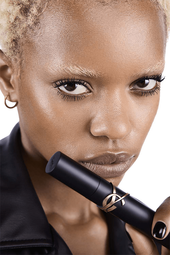 Une femme avec des cheveux blonds et courts tient un tube de mascara noir pres de son visage, affichant un regard intense. Elle porte une veste en cuir et des boucles d'oreilles discretes.