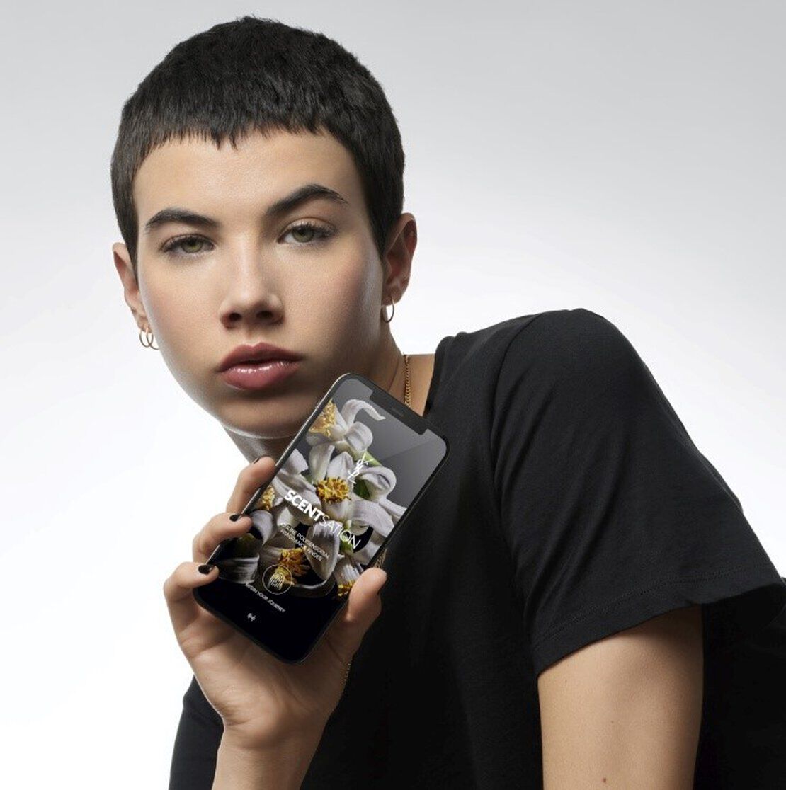 Une personne aux cheveux courts, portant un t-shirt noir, tient un smartphone avec un fond floral affiche a l'ecran.