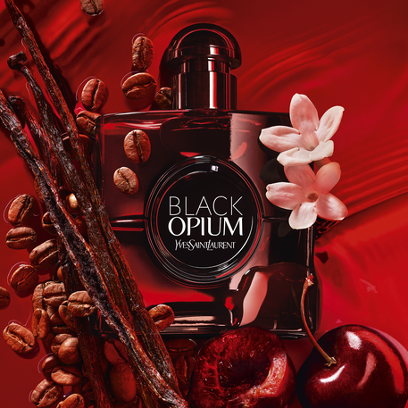 Flacon de parfum "Black Opium" d'Yves Saint Laurent, entoure de grains de cafe, de batons de vanille, de cerises et de fleurs blanches sur un fond rouge vibrant.