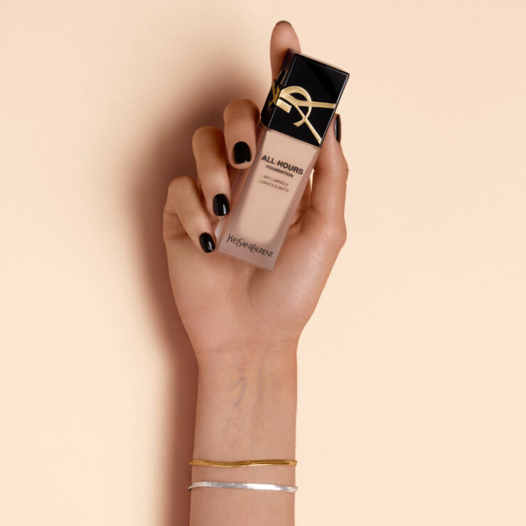 A hand holding a bottle of YSL All Hours foundation against a beige background. The bottle features a sleek design with a black cap and gold logo. The hand has dark nail polish and is adorned with simple bracelets.