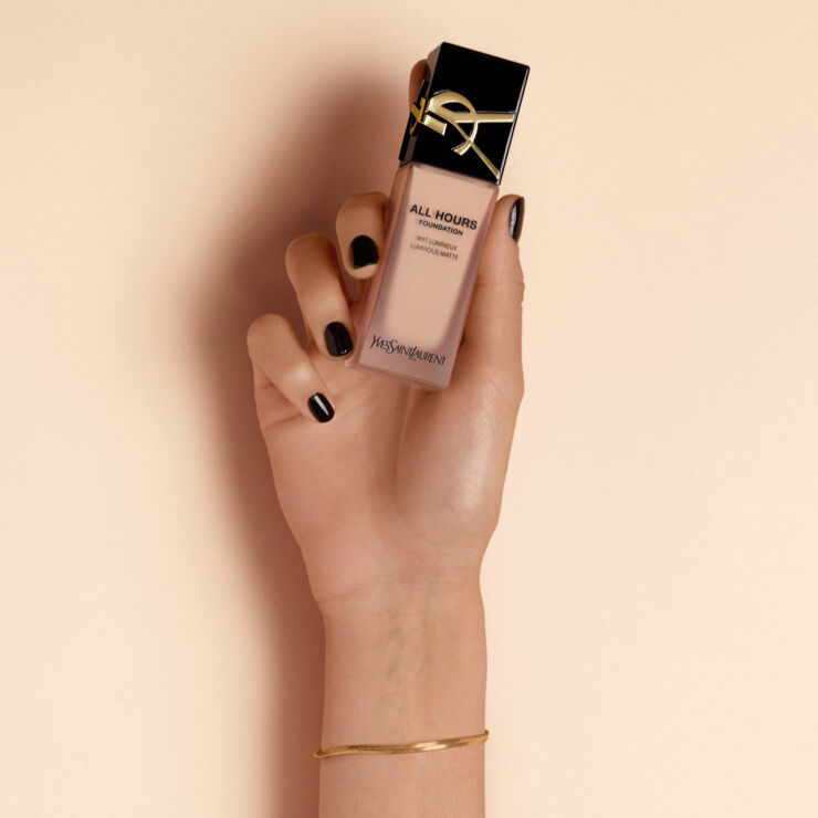 A hand with black nail polish holding a bottle of YSL All Hours foundation against a light peach background.