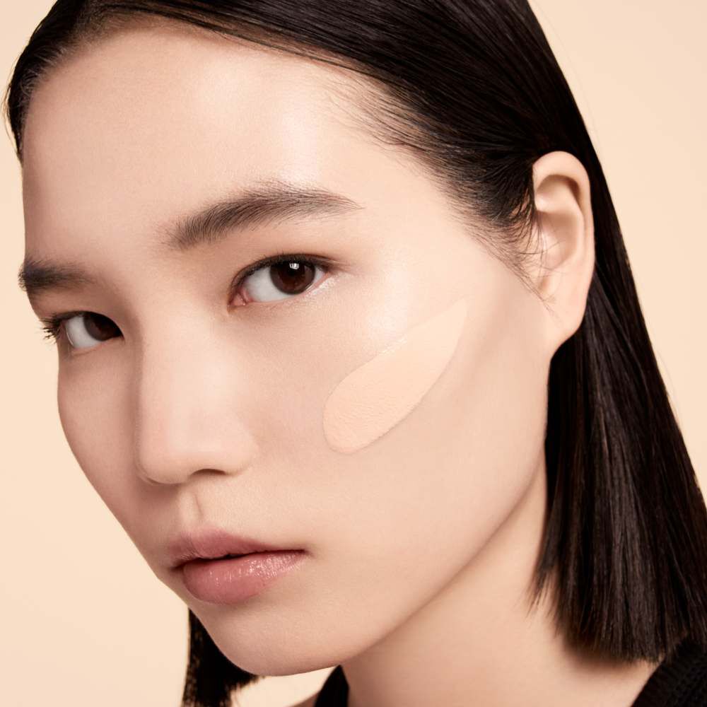 A close-up portrait of a young woman with straight black hair, applying a light beige foundation on her cheek. She has a neutral expression and a natural makeup look against a soft, neutral background.