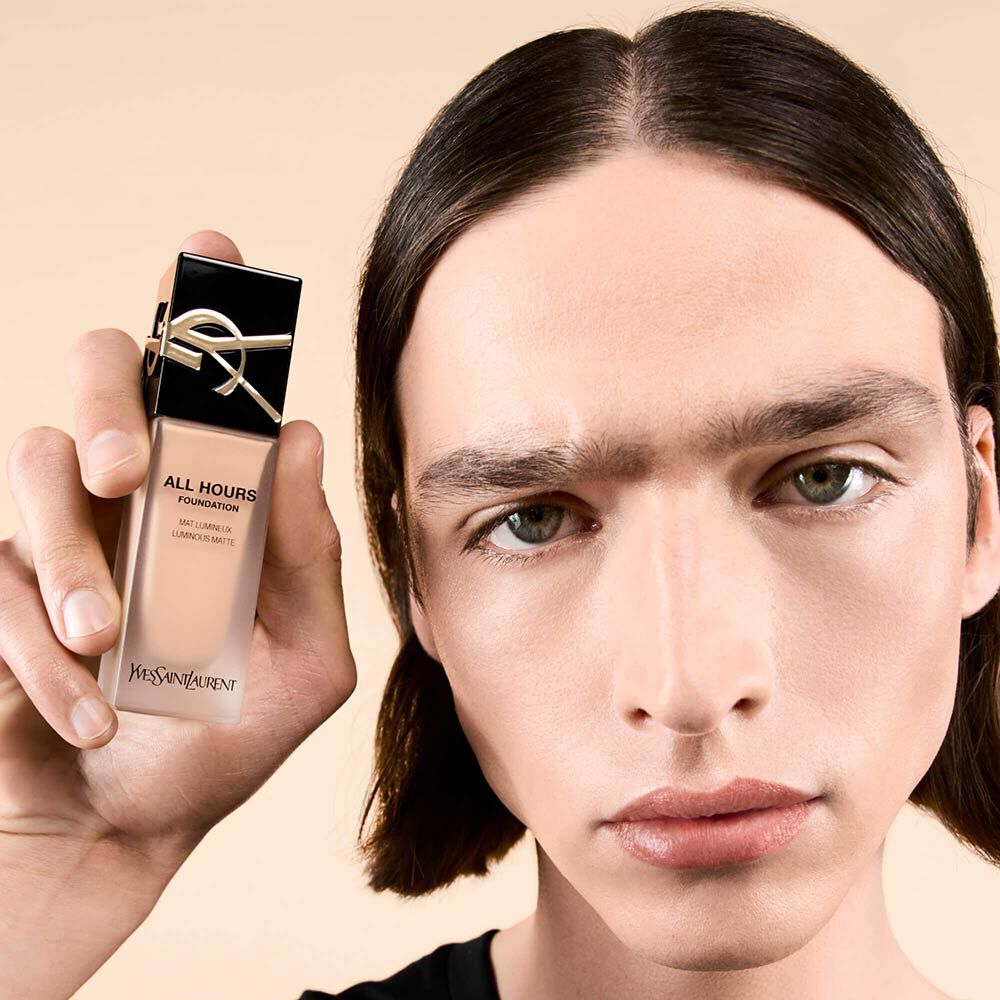 A close-up of a young person holding a bottle of YSL All Hours Foundation, showcasing their natural makeup look and serious expression against a neutral background.