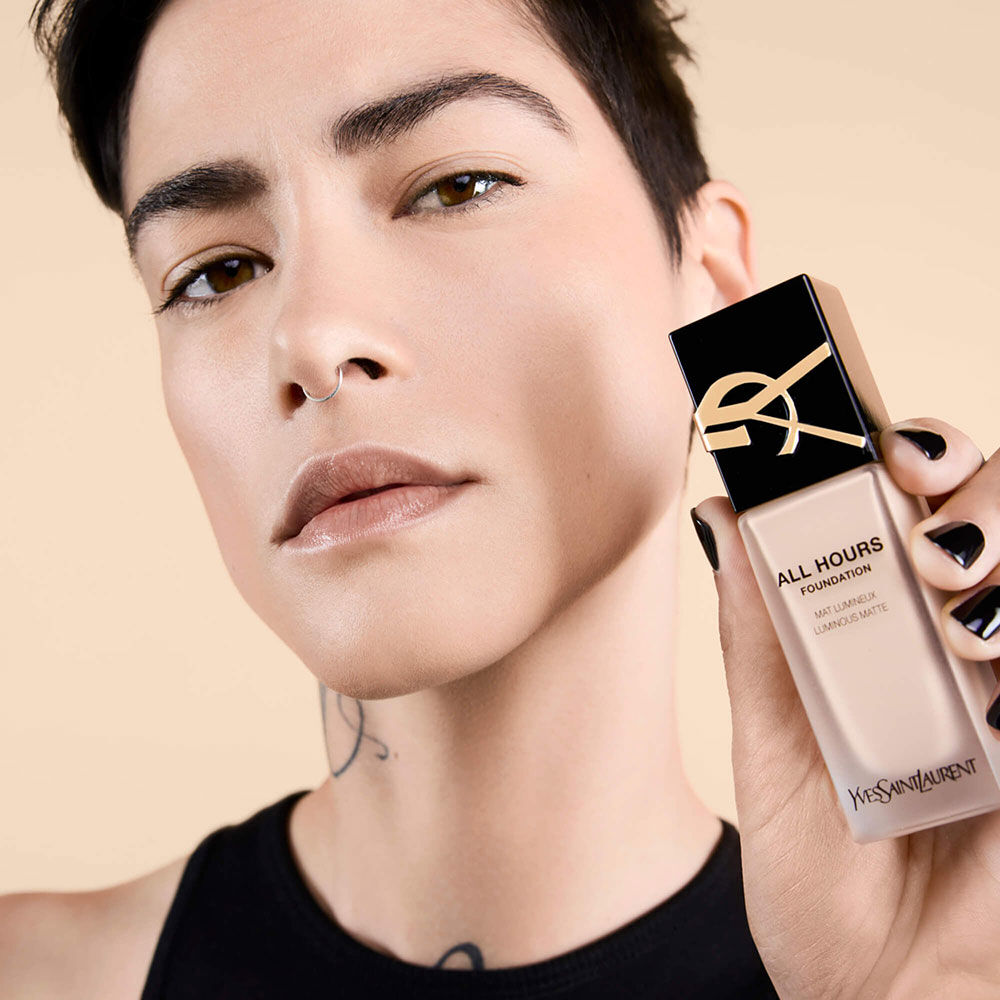 A close-up of a person holding a bottle of Yves Saint Laurent All Hours Foundation, showcasing their natural makeup look with a neutral background.