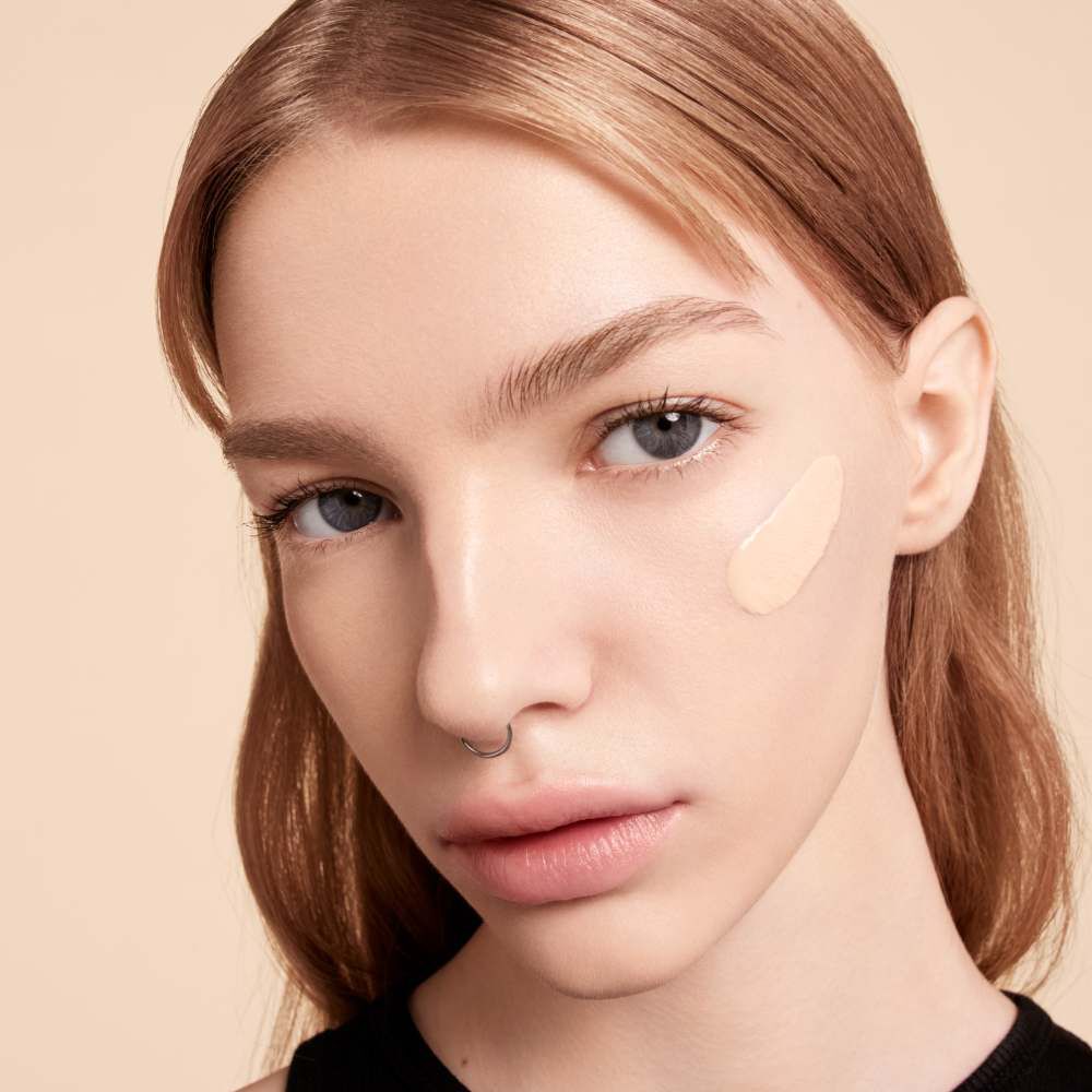 A close-up portrait of a model with light skin and straight blonde hair, showcasing a foundation swatch on her cheek. She has a septum piercing and is looking directly at the camera with a neutral expression.