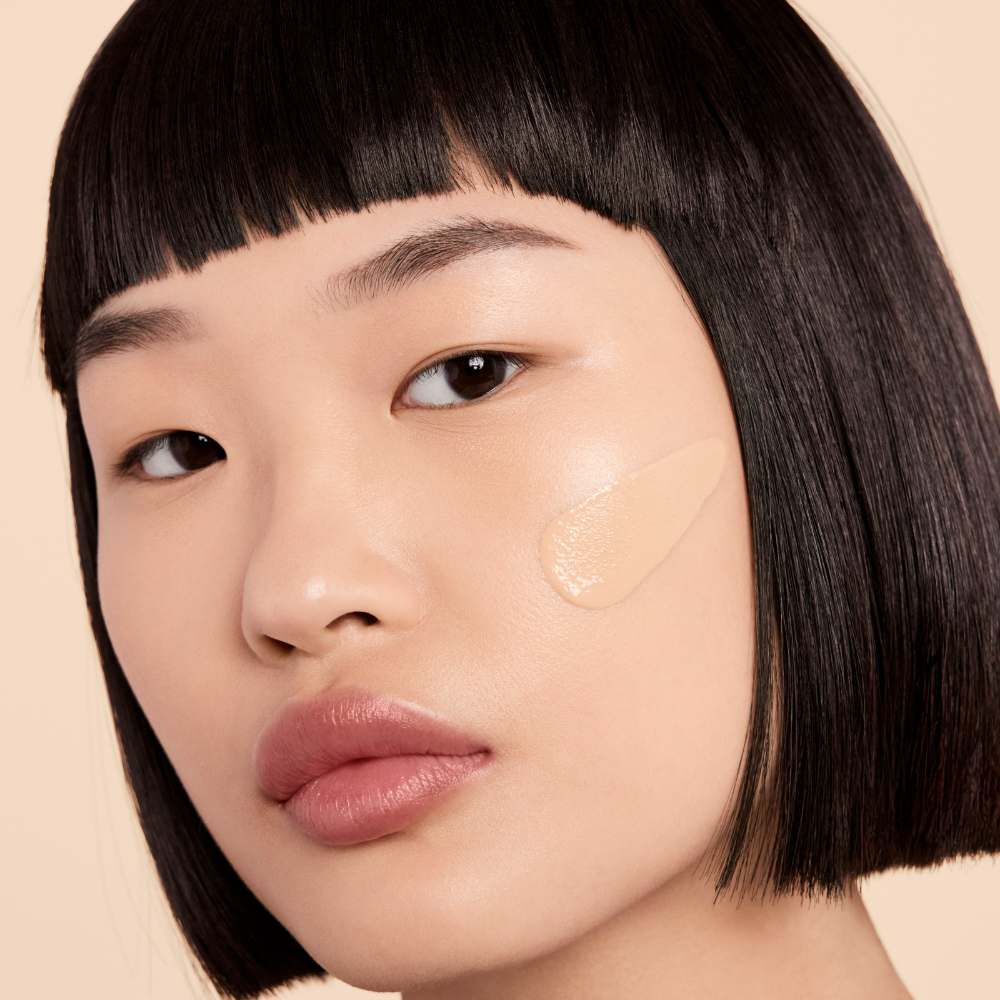 A close-up of a woman with straight black hair and a blunt bob hairstyle, showcasing a smooth complexion. She has foundation applied to her cheek, highlighting natural beauty.