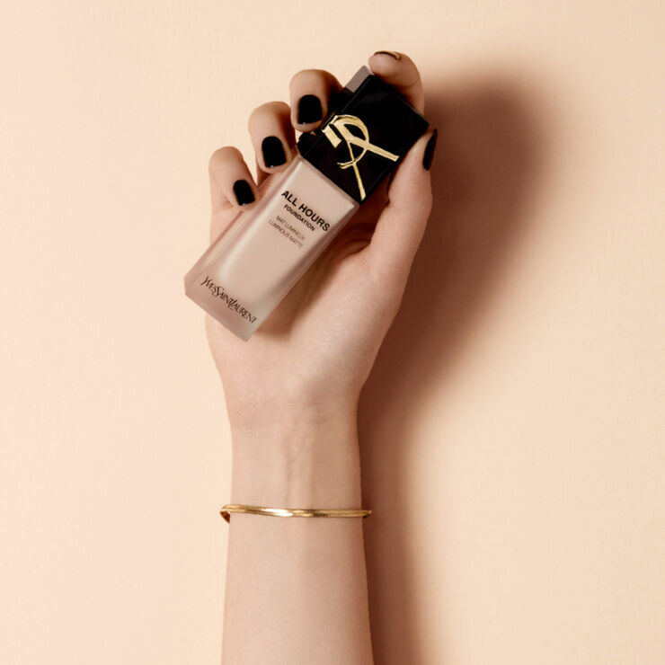 A hand with black nail polish holds a bottle of foundation from Yves Saint Laurent, featuring a sleek design and a gold ribbon on the cap, against a soft beige background.