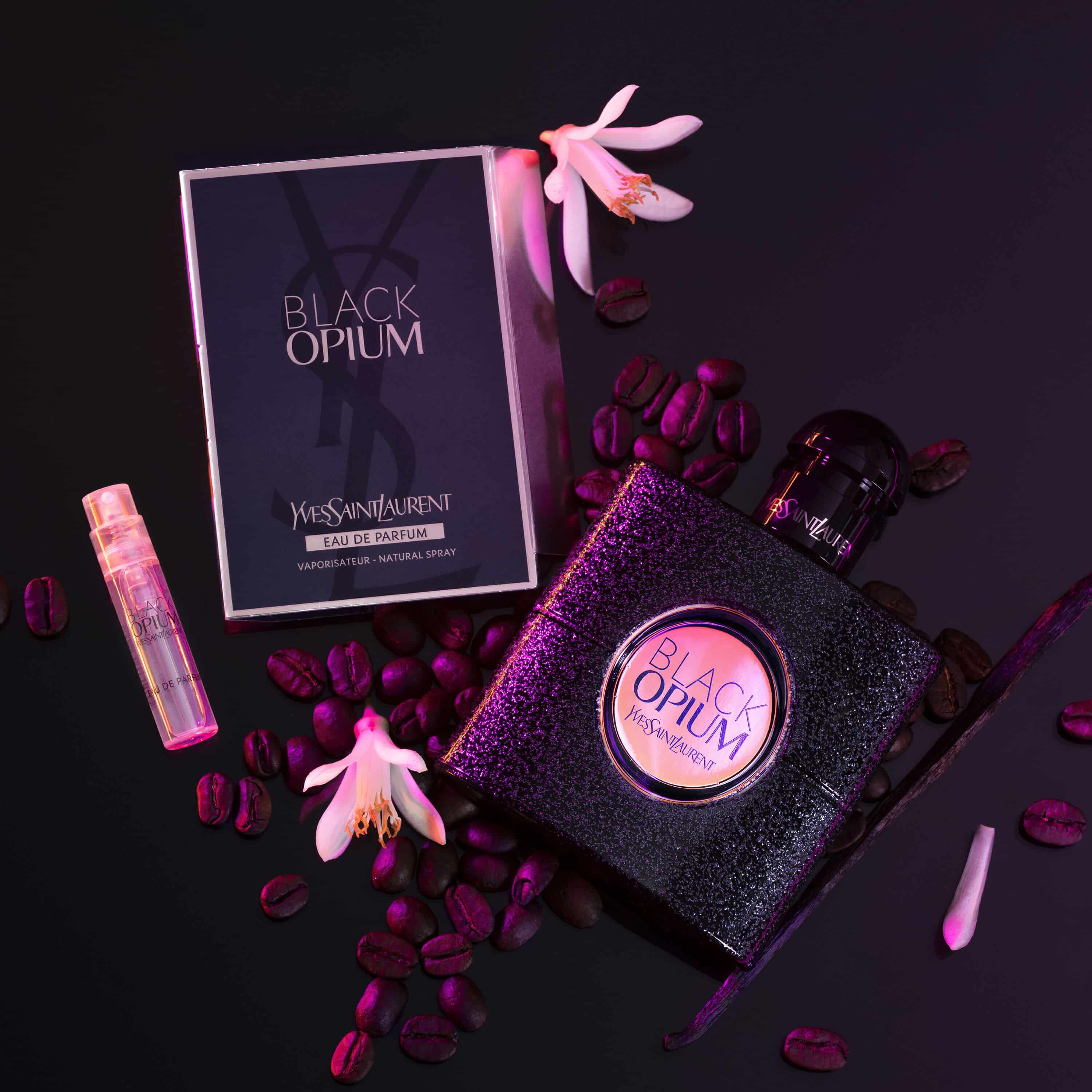 Flacon de parfum "Black Opium" d'Yves Saint Laurent, accompagne de sa boite et d'echantillons, entoure de grains de cafe et de fleurs blanches.