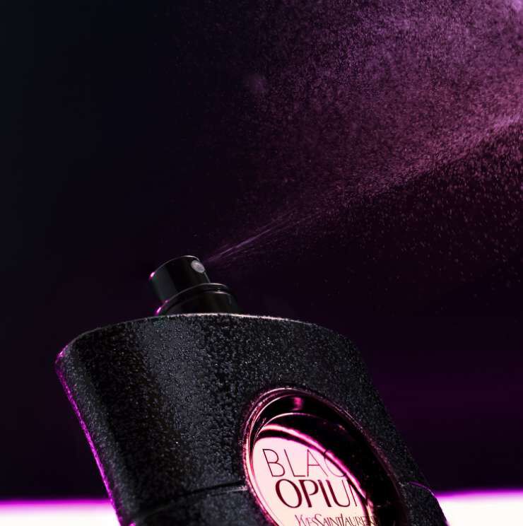 Un flacon de parfum noir avec un spray en train de diffuser une brume legere, sur un fond sombre.