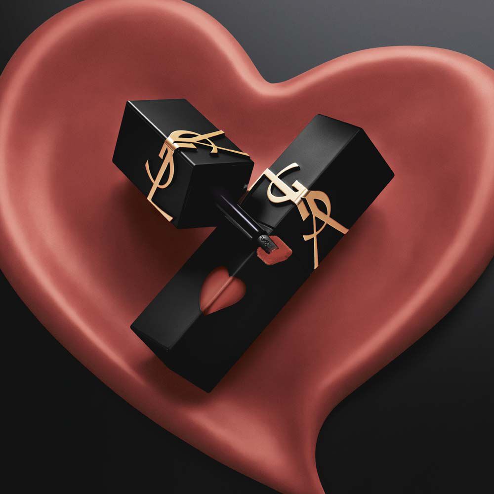 Deux mannequins chic en trench noir et veste de cuir, tenant des rouges a levres YSL devant un immense fond rose en forme de coeur. Une bouche rouge brillante, une bouche rose mat veloute.