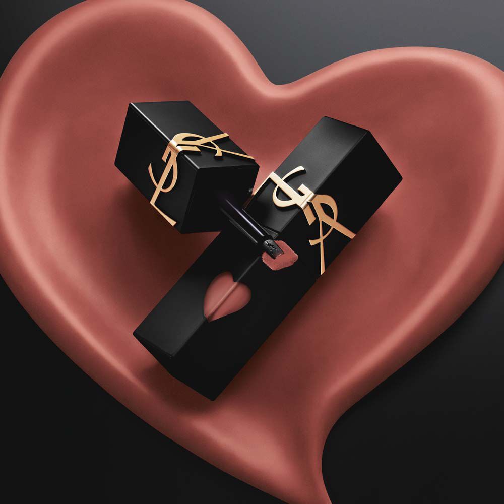 Deux flacons de teinture a levres YSL mat noir aux decoupes en forme de coeur rose et logos dores, exposes dans un coeur sculpte mat rose assorti sur fond noir.