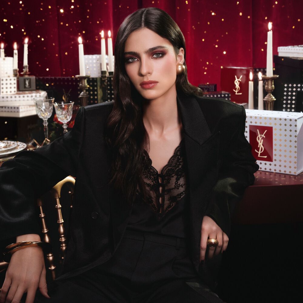 Mannequin en costume noir et look glamour, assise dans un decor festif baigne de bougies, entouree de coffrets cadeaux YSL.