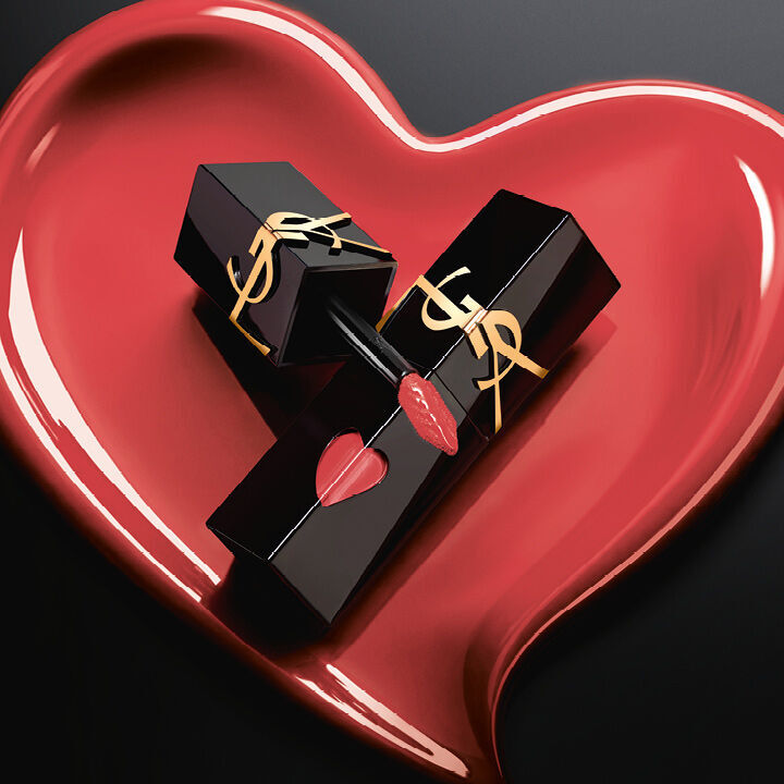 Deux flacons de teinture a levres YSL noir mat aux decoupes en forme de coeur rose et logos dores, exposes dans un coeur sculpte rose mat assorti sur fond noir.