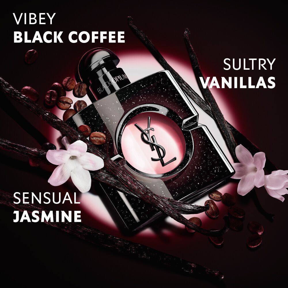 Gros plan sur des grains de cafe torrefies, des gousses de vanille et des fleurs de jasmin disposes autour de l’iconique flacon Black Opium Eau de Parfum orne de la Cassandre YSL.