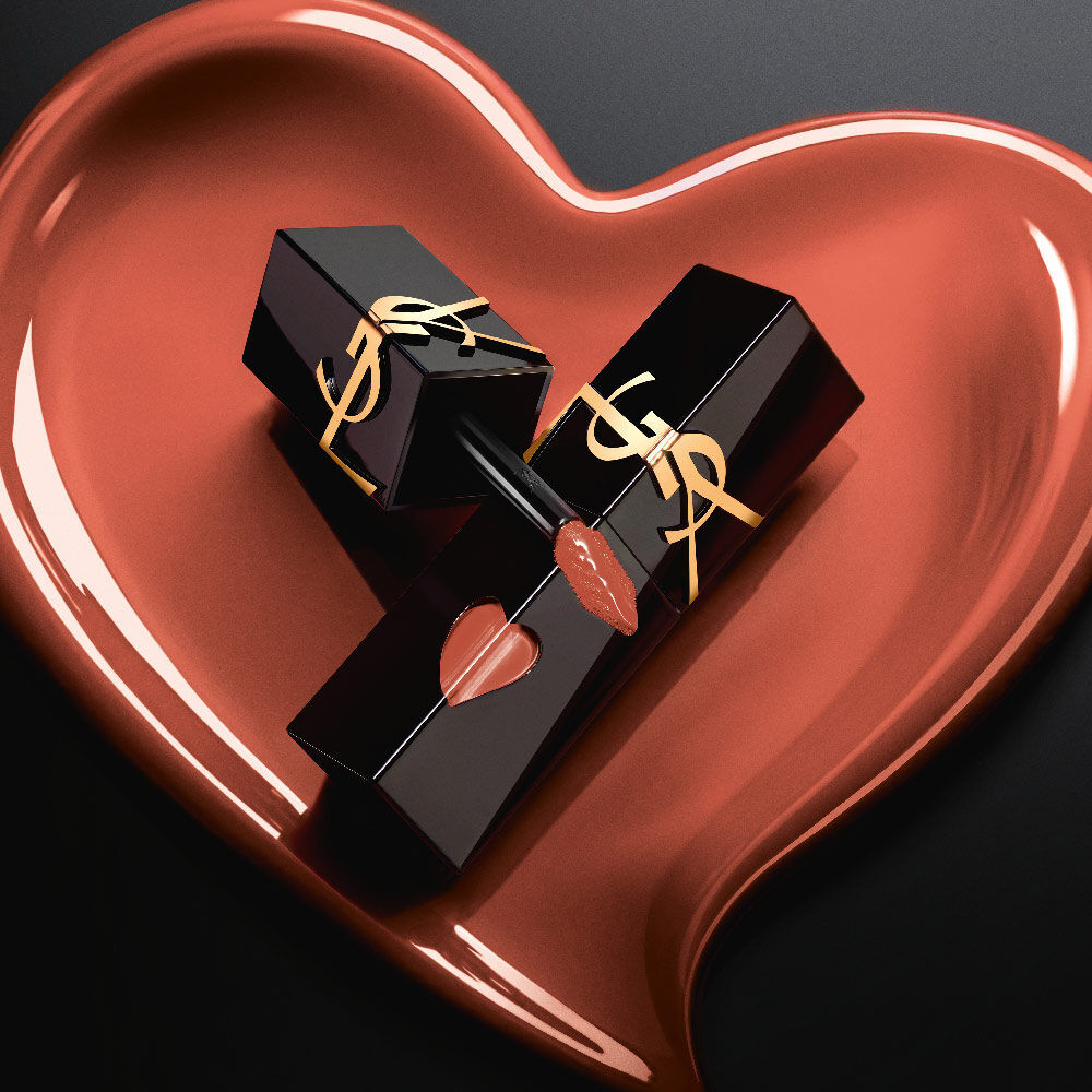 Deux flacons de teinture a levres YSL noir mat aux decoupes en forme de coeur rose et logos dores, exposes dans un coeur sculpte rose mat assorti sur fond noir.