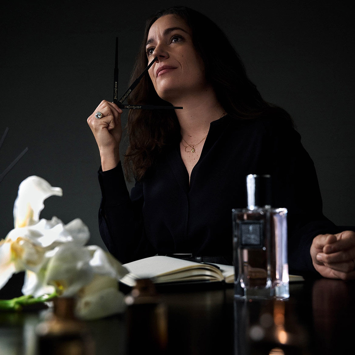 Une femme en chemise noire tient une touche à parfum, levant les yeux pensivement, avec un carnet, une bouteille de parfum et des fleurs sur la table.