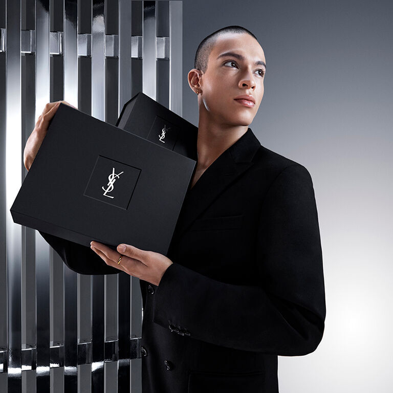 Pierre-Antoine, un mannequin homme, tient avec élégance un coffret cadeau YSL Beauty noir arborant l'iconique logo Cassandre.