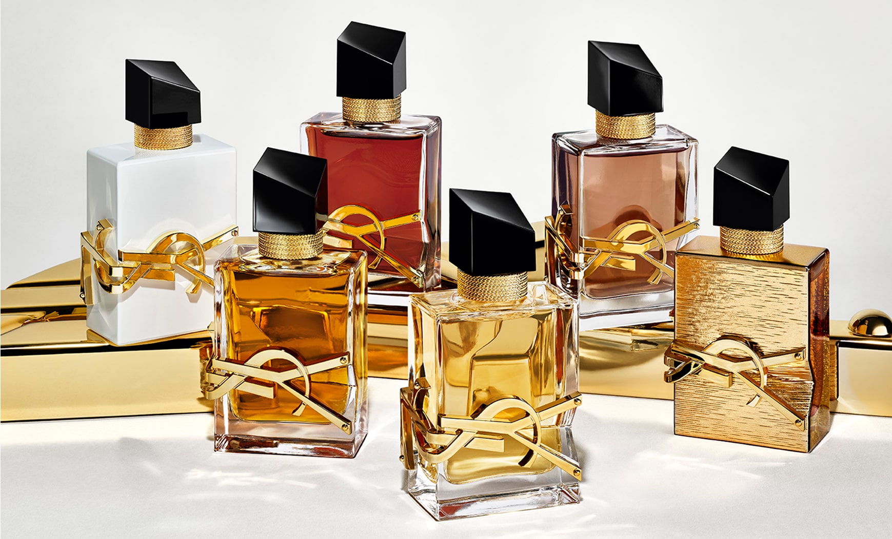 Six flacons de parfum YSL Libre sont présentés sur une surface dorée brillante. Les flacons présentent des variations de couleur du liquide et de finition : transparent, blanc, rougeâtre et brun, ainsi qu'un flacon texturé entièrement doré. Tous arborent l'emblématique lettrage YSL doré et un bouchon noir incliné.
