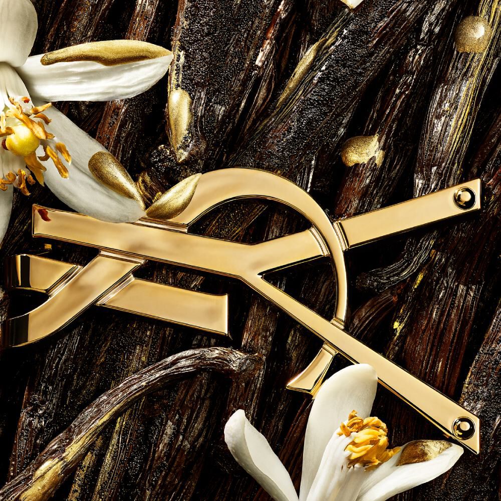 Gros plan du logo YSL doré posé parmi des gousses de vanille, des fleurs blanches et des touches dorées scintillantes.