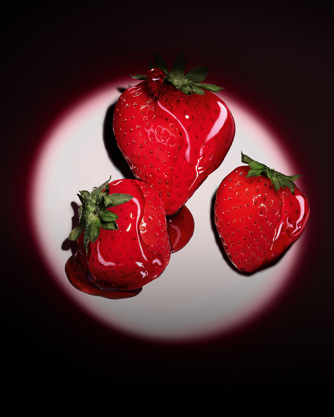 Trois fraises rouges fraîches illuminées par un projecteur circulaire rose sur un fond sombre.