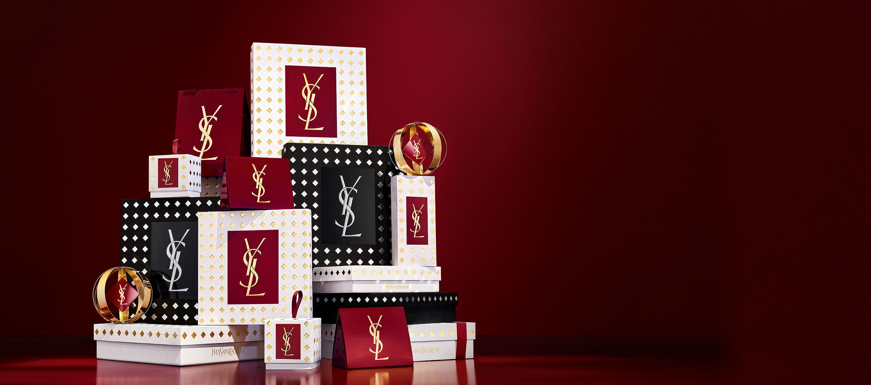 Trois mannequins en tenues noires élégantes tenant des coffrets cadeaux YSL des fêtes devant un rideau rouge scintillant, avec des confettis dorés qui tombent.