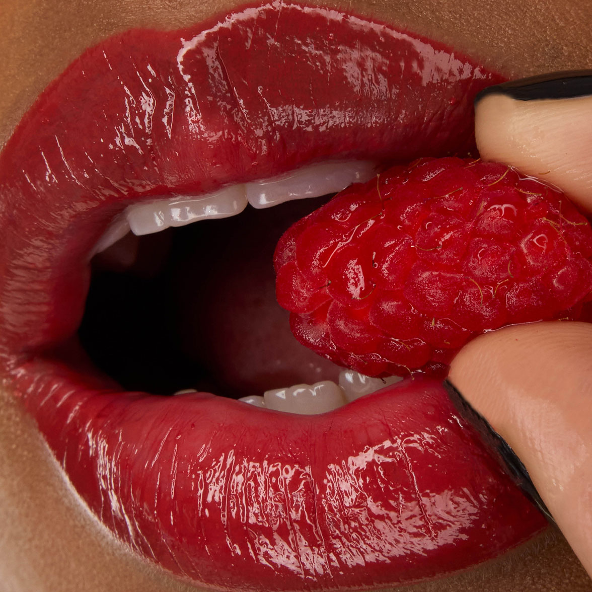 Gros plan sur les lèvres du mannequin Zahara, habillées du rouge à lèvres YSL LOVESHINE en teinte Berry Crush, nuance 219, tenant une framboise fraîche.