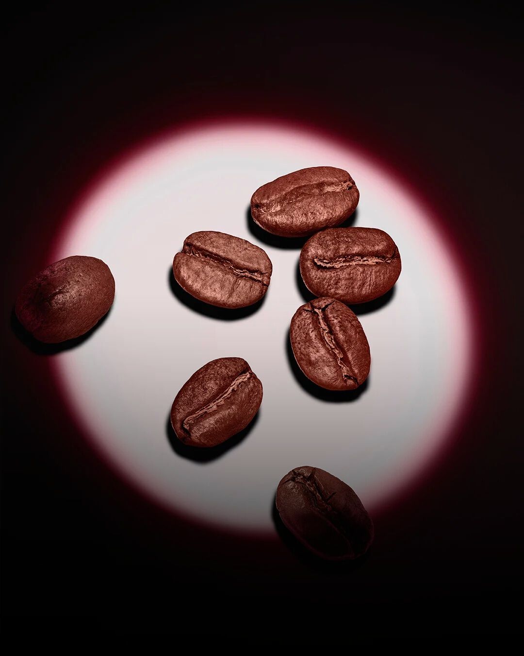 Un groupe de grains de café torréfiés, mis en lumière par un halo circulaire rose sur fond sombre.