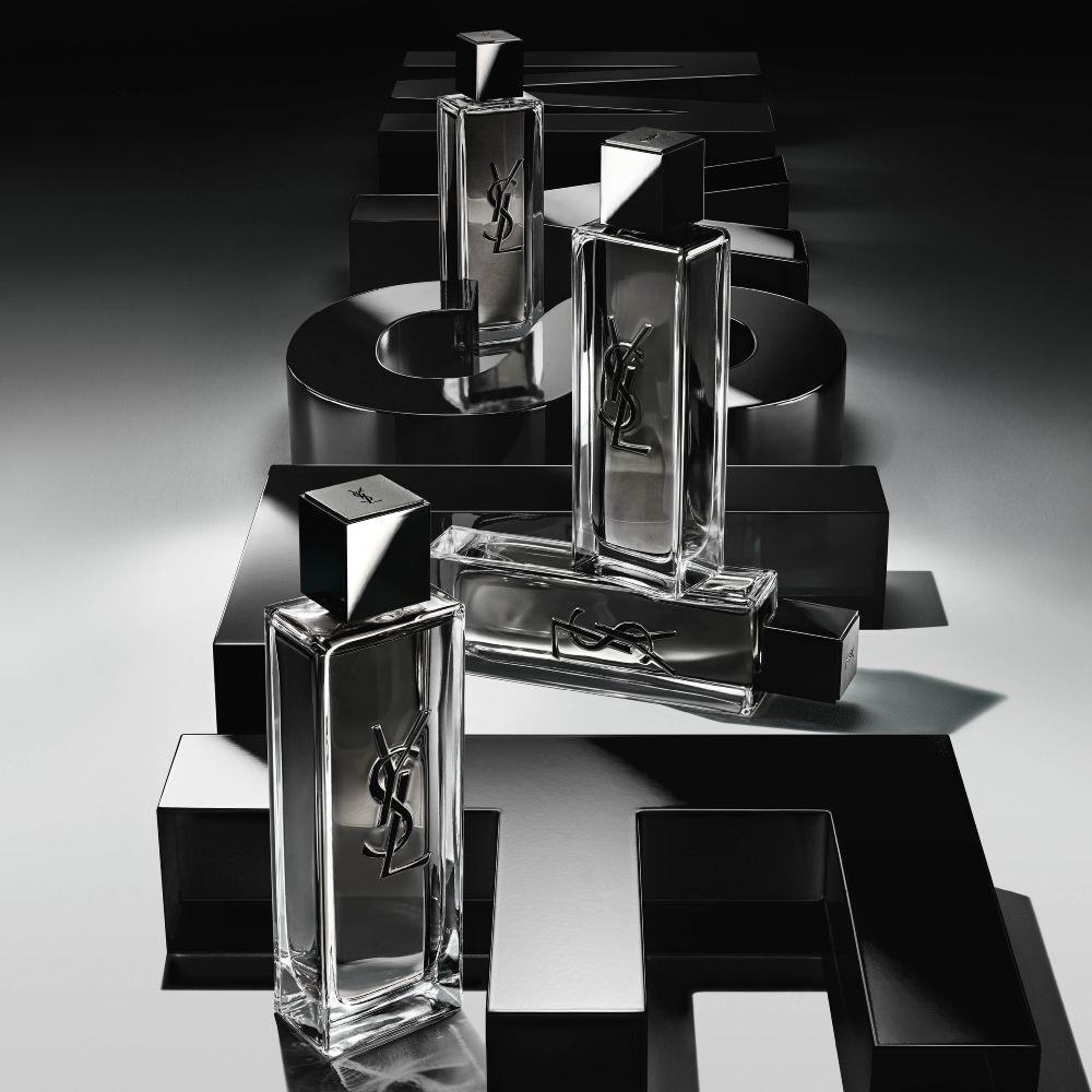 Vue en plongée de flacons de MYSLF Eau de Toilette Intense disposés sur de grandes lettres 3D noires formant le mot MYSLF, posées à plat sur le sol.