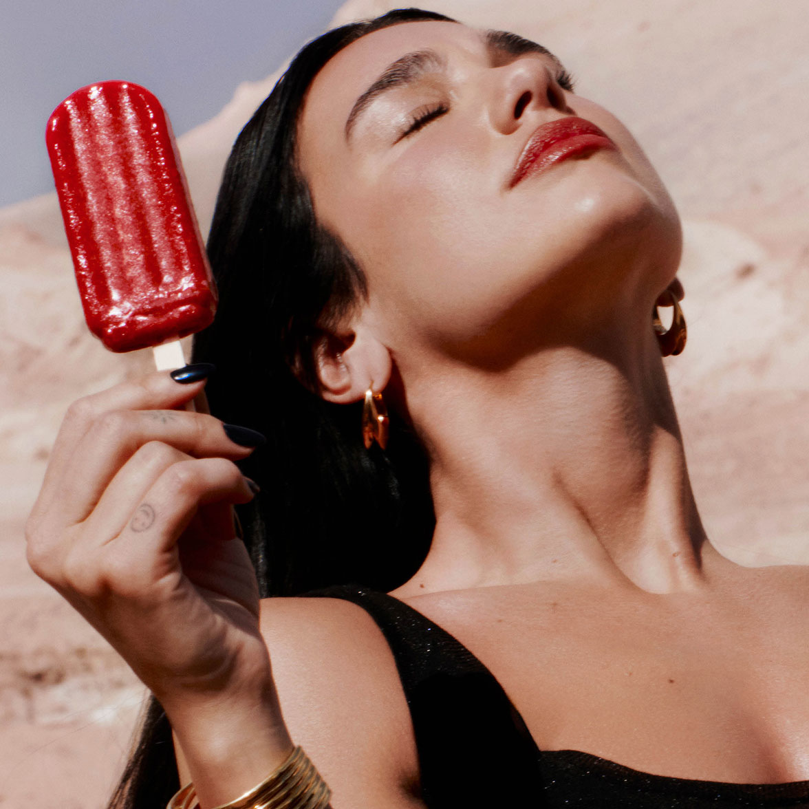 Un gros plan de Dua Lipa, les yeux fermés au soleil, tenant une glace à l'eau rouge.