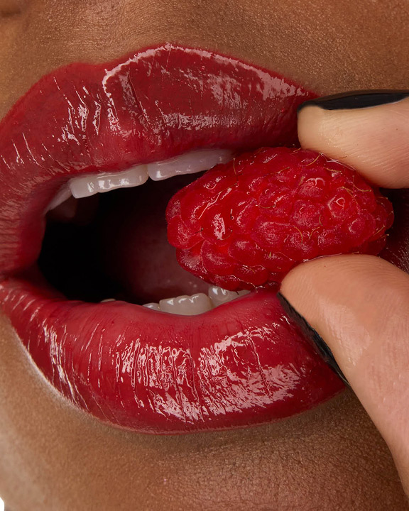 Gros plan sur les lèvres du mannequin Zahara, habillées du rouge à lèvres YSL LOVESHINE en teinte Berry Crush, nuance 219, tenant une framboise fraîche.