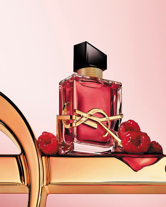 Flacon de parfum YSL rouge avec logo doré, sur une surface dorée avec des framboises.
