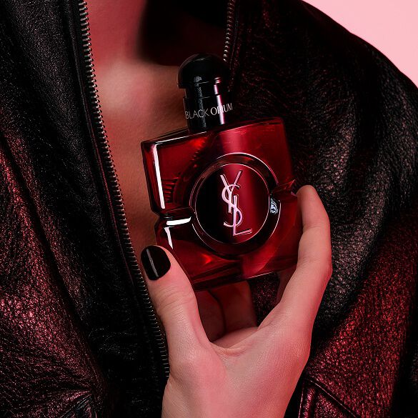 Gros plan d’une main aux ongles noirs tenant le flacon Black Opium Eau de Parfum Over Red sur fond de veste en cuir noir.