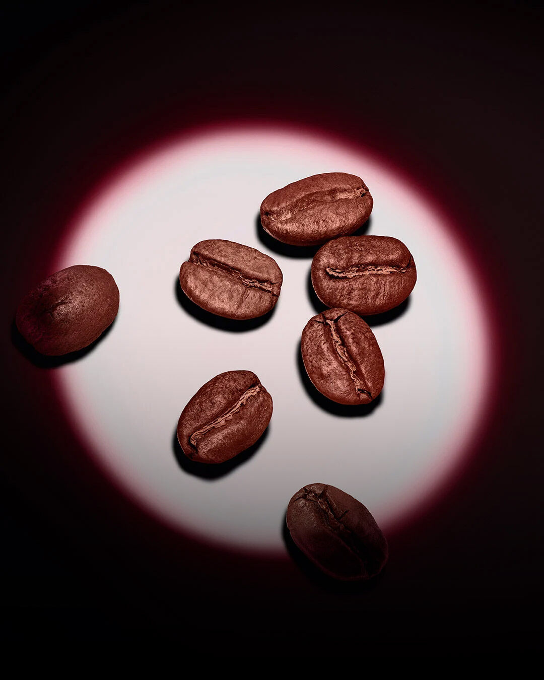 Un groupe de grains de café torréfiés illuminés par un projecteur circulaire rose sur un fond sombre.