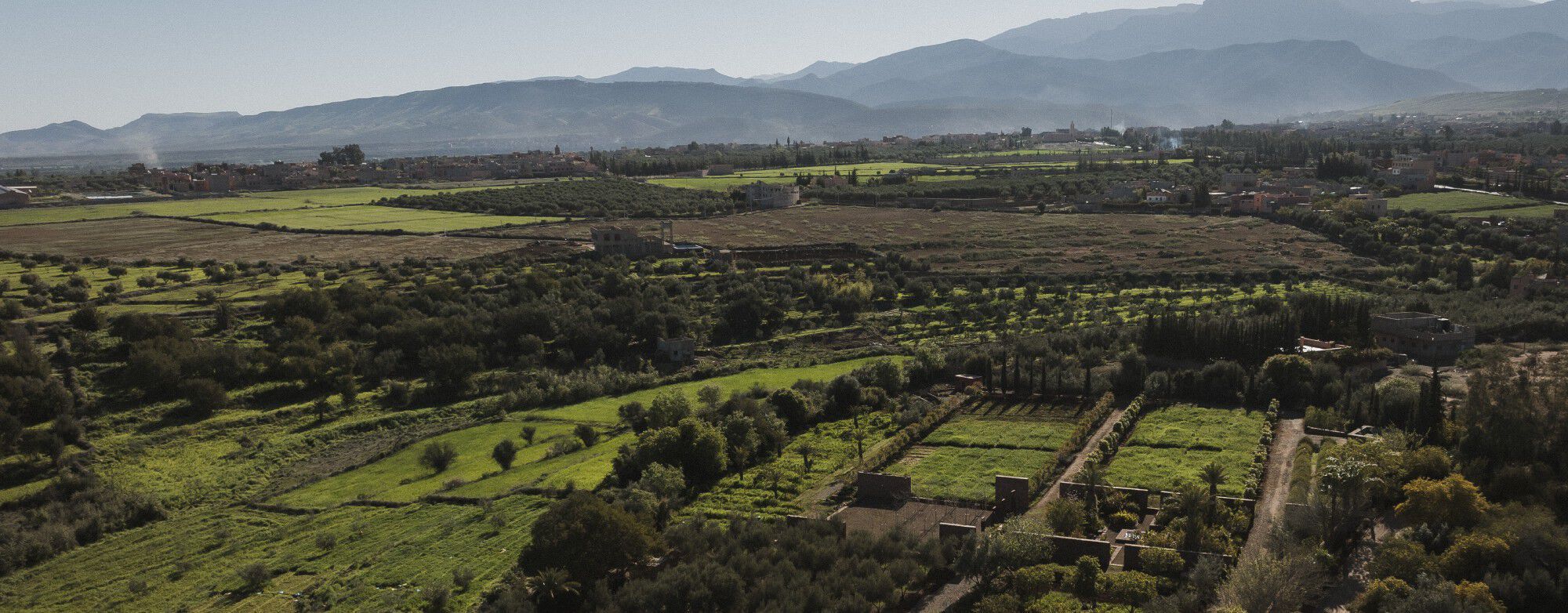 YSL For The Future : Vue aérienne des Jardins Collectifs de l'Ourika, avec des champs verdoyants, des arbres et des montagnes sous un ciel dégagé.